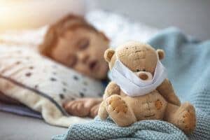 Little boy sleeping in bed with medical mask embracing his teddy bear. Home isolation coronavirus covid-2019 quarantine.